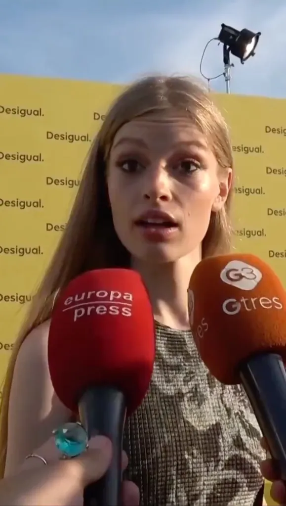 A young woman with long light brown hair is being interviewed by two microphones from Europa Press and Gtres, in front of a yellow banner with the word "Desigual" repeated.