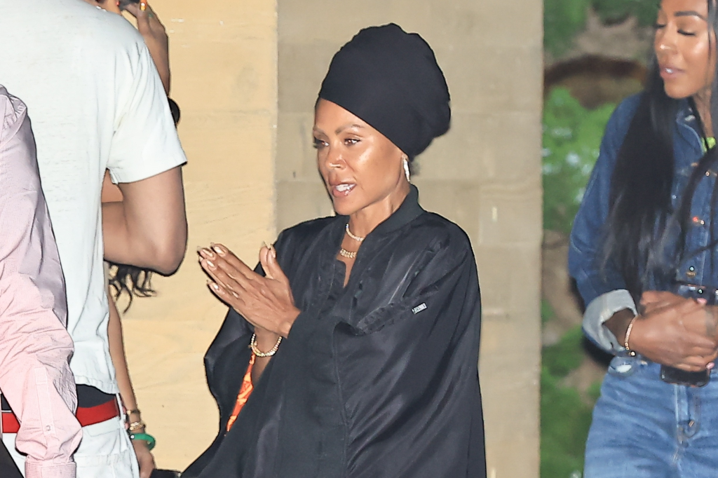 Jada Pinkett Smith speaking, wearing a black turban, black jacket, and gold necklaces, with two other people partially visible.