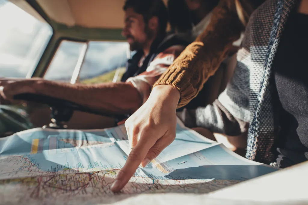 A person's hand pointing at a map on a road trip, with the driver in the background.