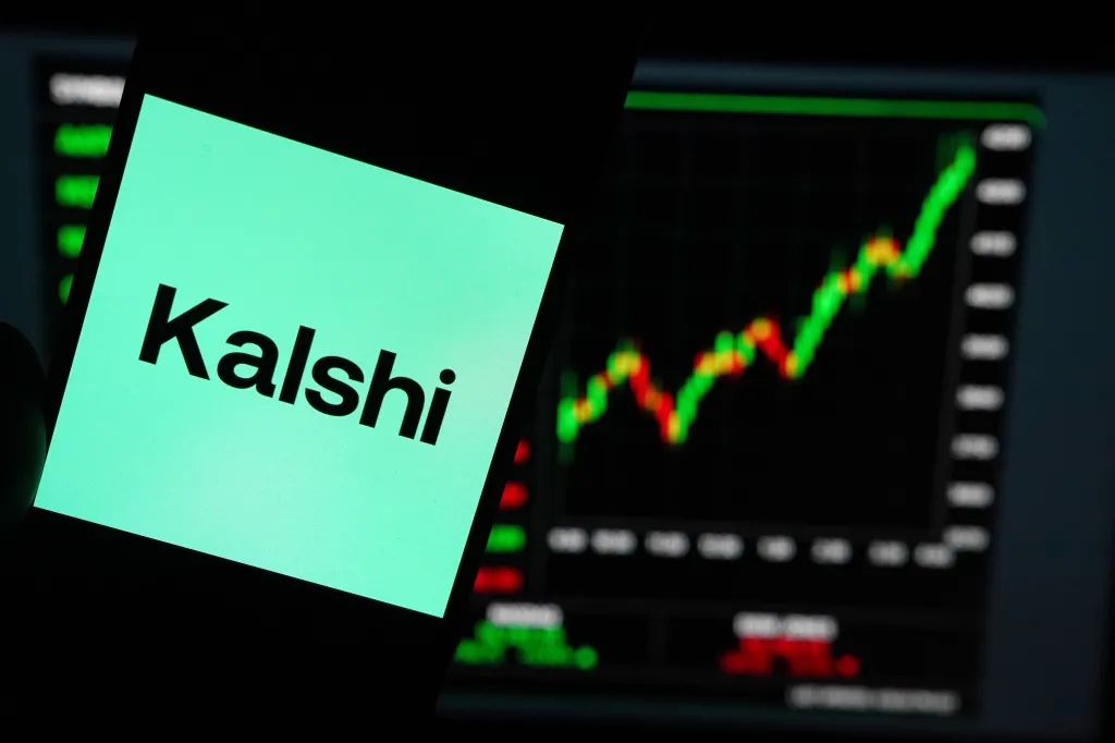 A smartphone displaying the Kalshi logo in front of a stock market chart with green and red lines.