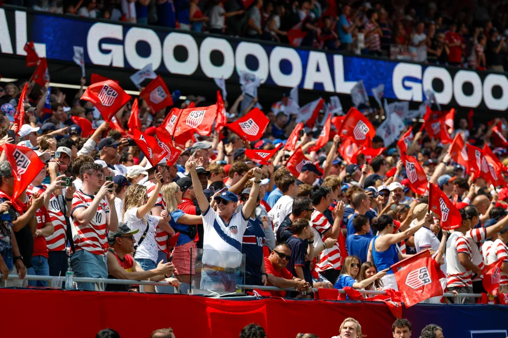 USA soccer fans celebrating a goal, holding red flags with "USA" printed on them, with a screen displaying "GOOOOOAL" in the background.
