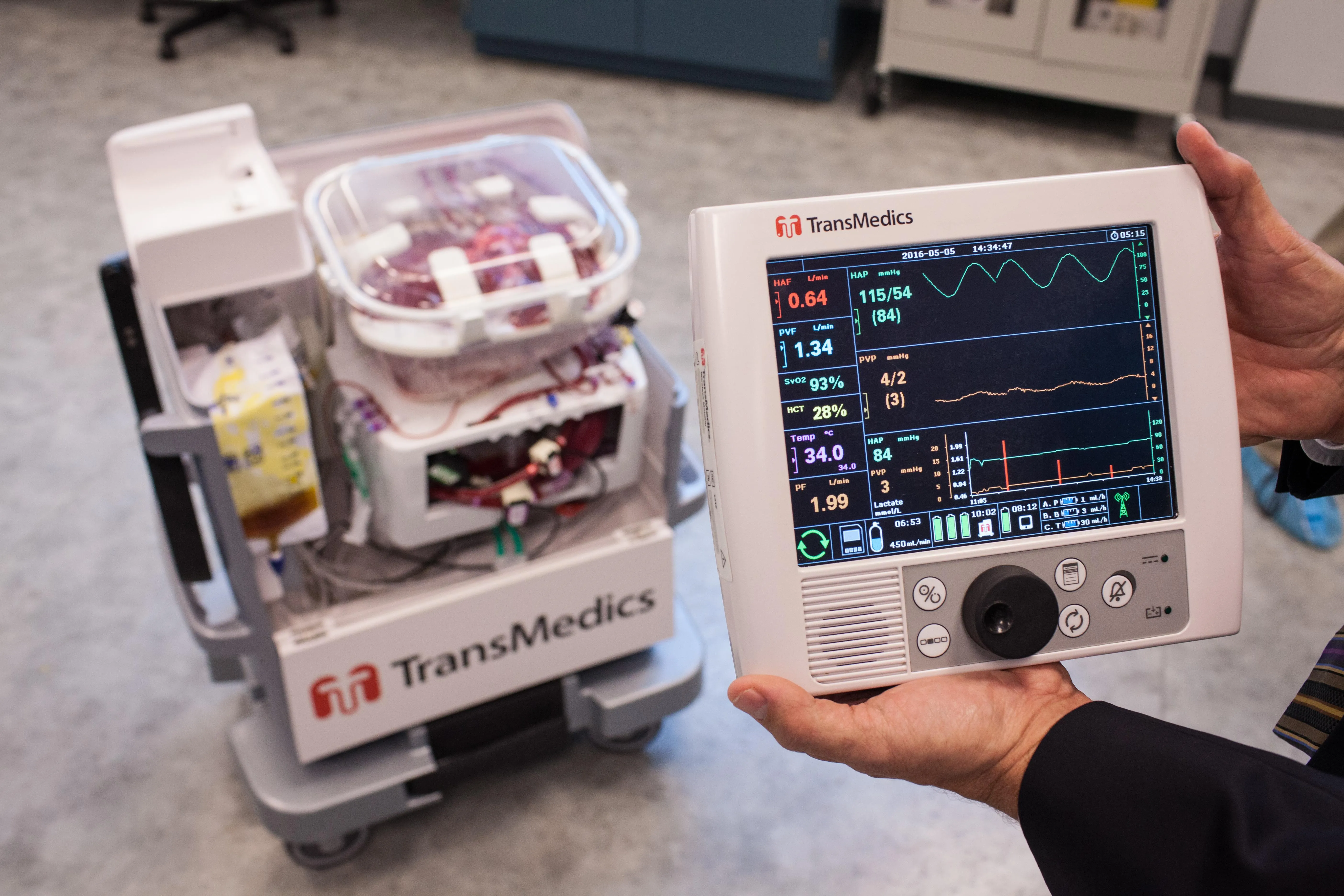 A man’s hands hold an electronic medical device in the foreground while a robotic cart sits in the background A man’s hands hold an electronic medical device in the foreground while a robotic cart sits in the background