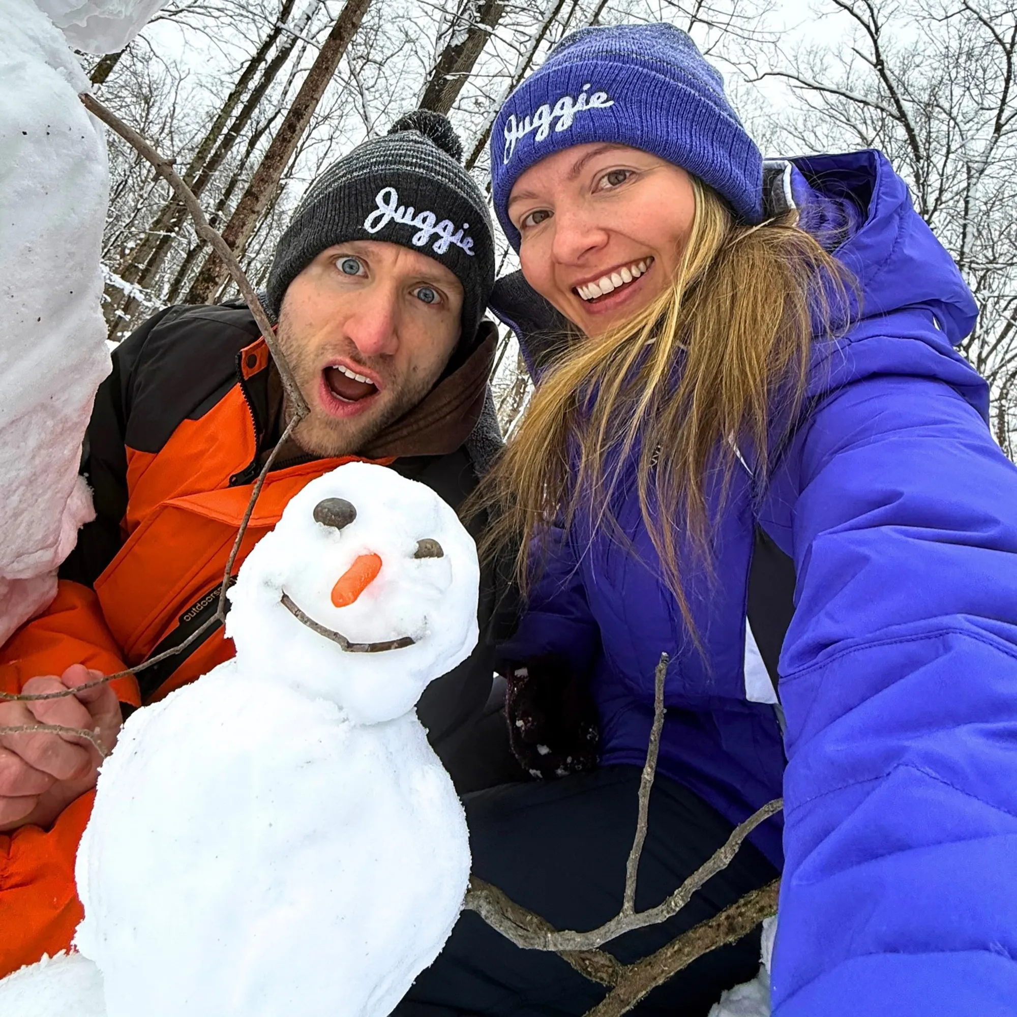 Jesse Ridgway and his wife, wearing beanies that say "Juggie," pose with a small snowman in a snowy, wooded area.