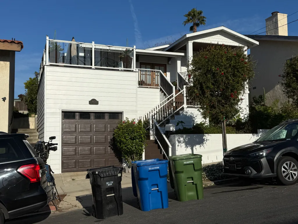 A white house with a brown garage door, front steps, and a second-story balcony. Three trash cans are on the street in front of the house.