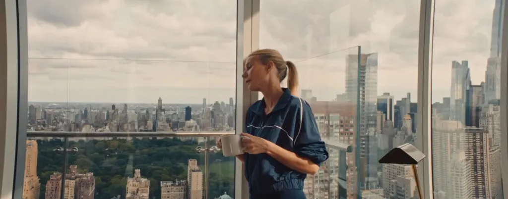 Gwyneth Paltrow, in a blue jacket, holding a coffee cup, looking out a window at a city skyline.