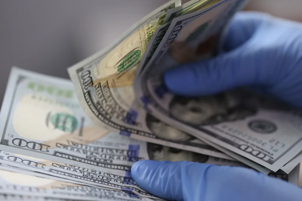 Hands in blue gloves holding one hundred dollar bills.