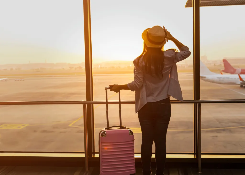 Woman at an airport holding a pink suitcase and looking at planes on the runway at sunset.