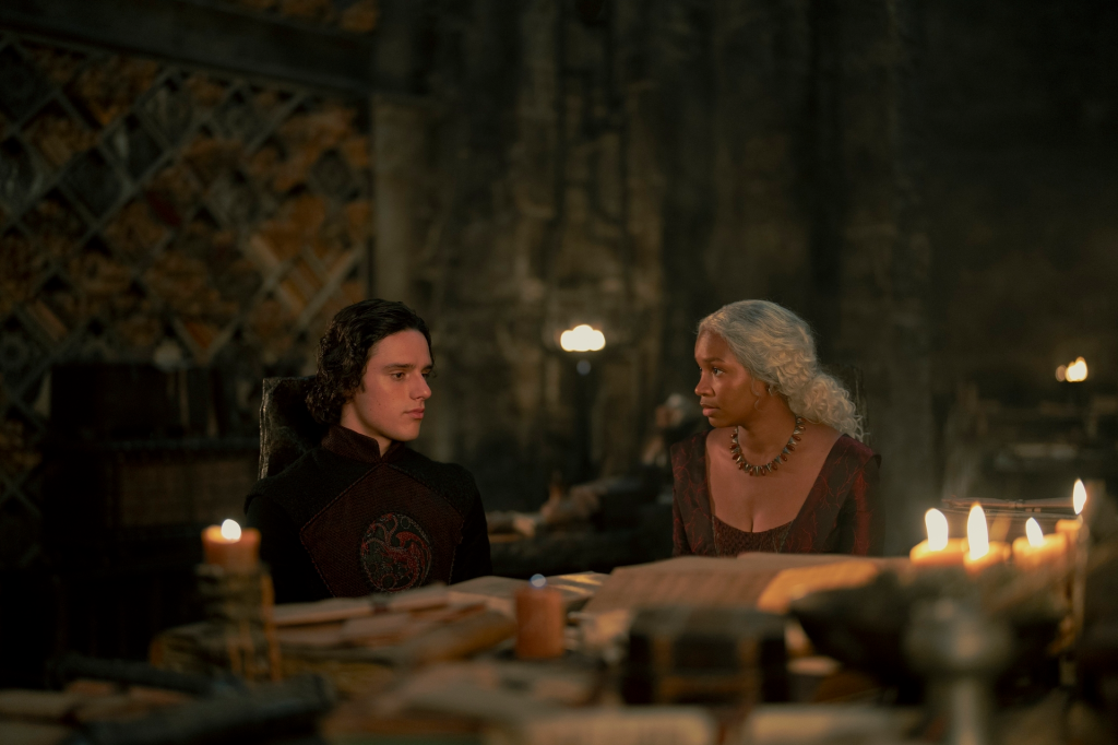 Harry Collett as Jacaerys Velaryon and an unnamed woman sitting at a table with books and candles in "House of the Dragon" Season 2.