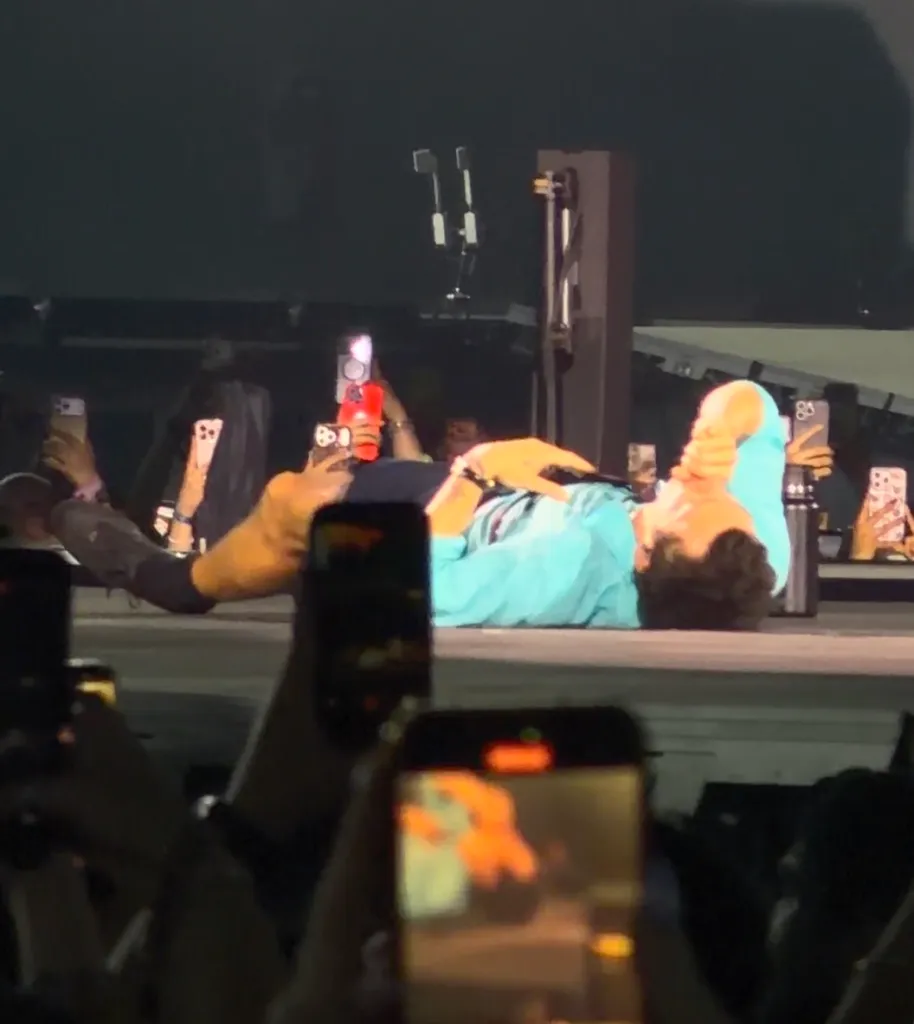 Harry Styles lies on a stage, surrounded by people holding up cell phones to record him.