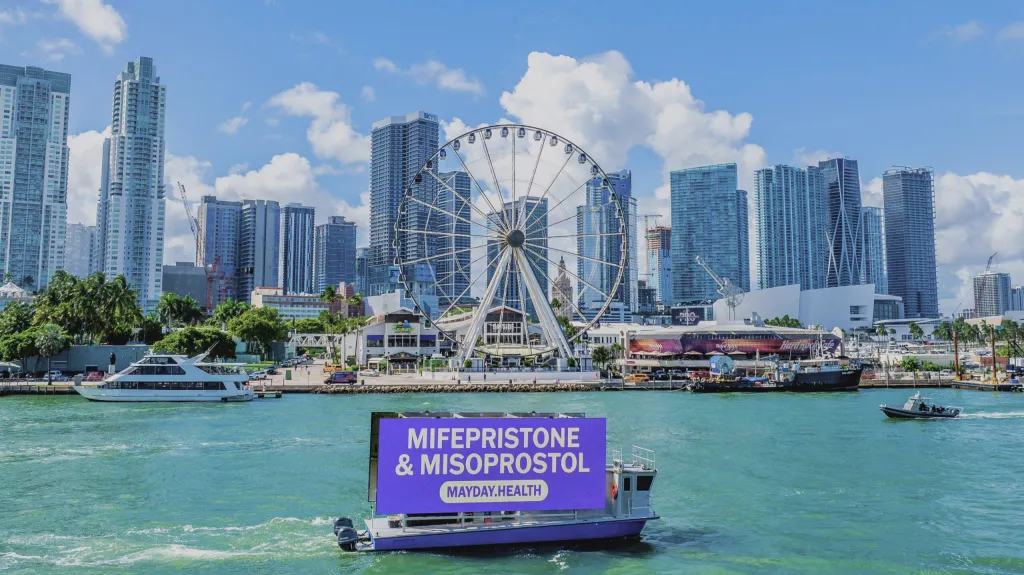A boat with a banner for Mayday Health advertising mifepristone and misoprostol in front of a city skyline.