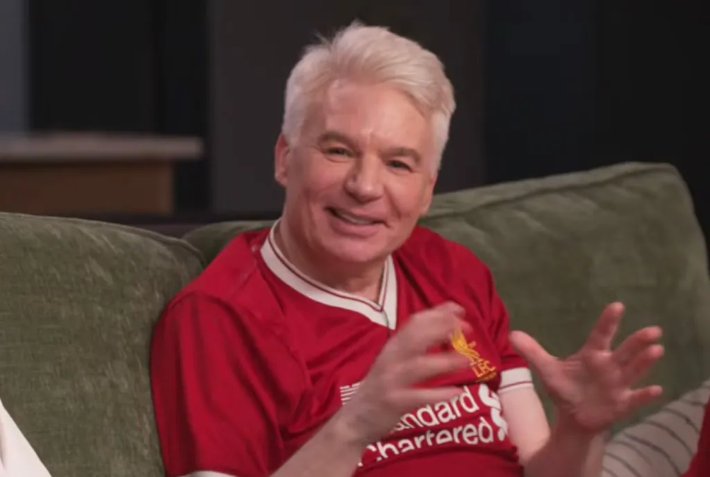 A light-skinned man with blond hair and a red Liverpool FC jersey smiles while gesturing with his hands.