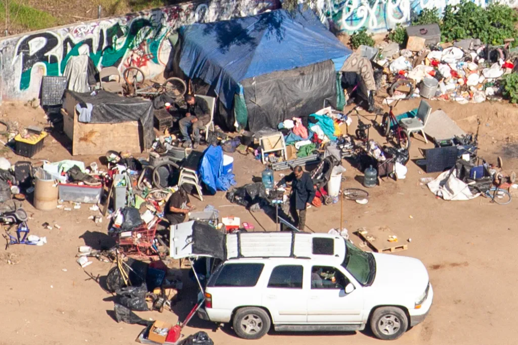 Aerial view of a homeless encampment with multiple tents, debris, and people, next to a graffiti-covered wall.