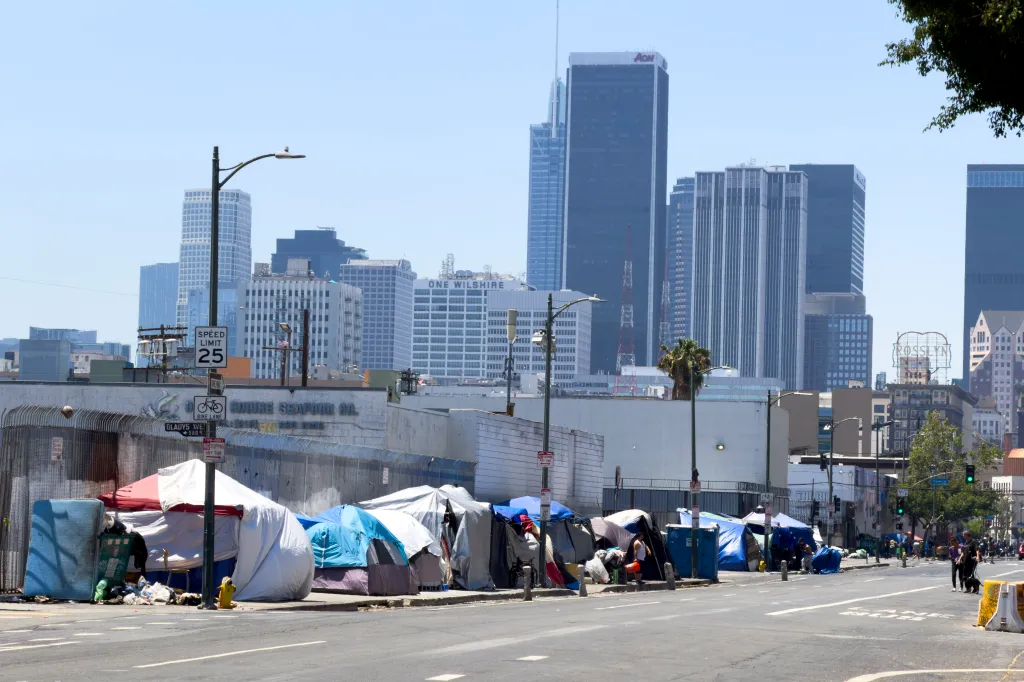 Homeless people and their tents lining a street in Skid Row, Los Angeles, with downtown buildings in the background.