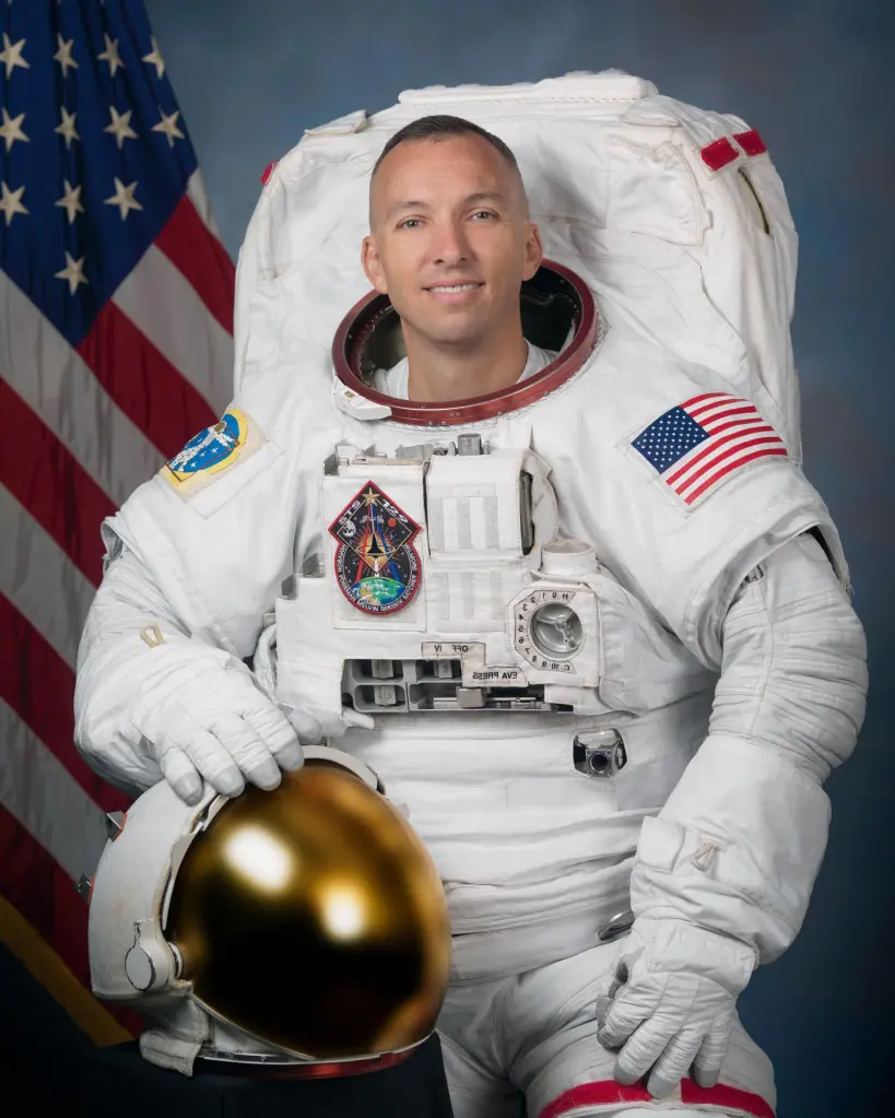 Astronaut Randy "Komrade" Bresnik in a white spacesuit with an American flag and a mission patch, holding a gold-visored helmet.