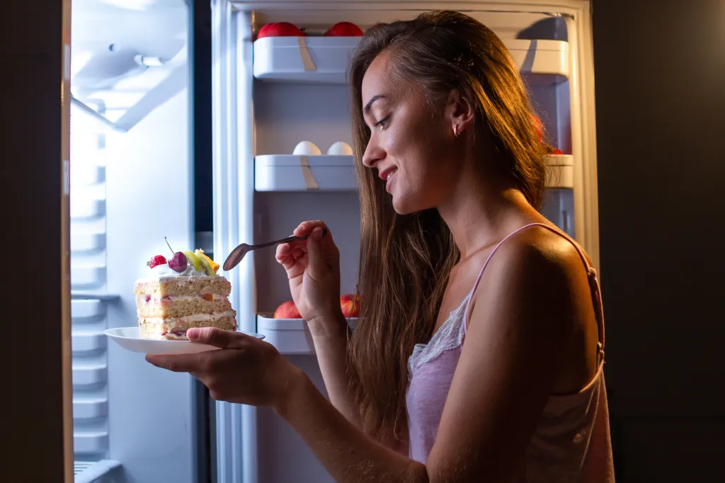 A woman in pajamas eating a slice of cake in front of an open refrigerator at night.