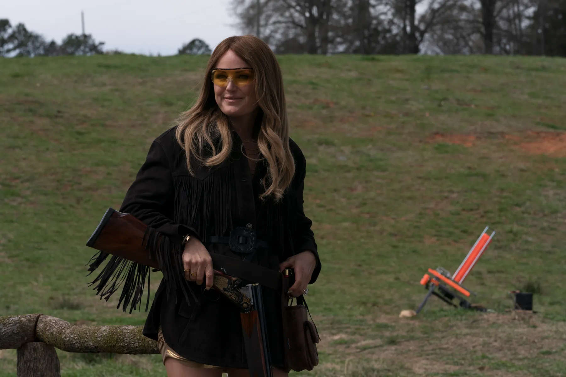 Margo Banks (Malin Åkerman) holding a shotgun, wearing a black fringed jacket, gold shorts, and yellow glasses.