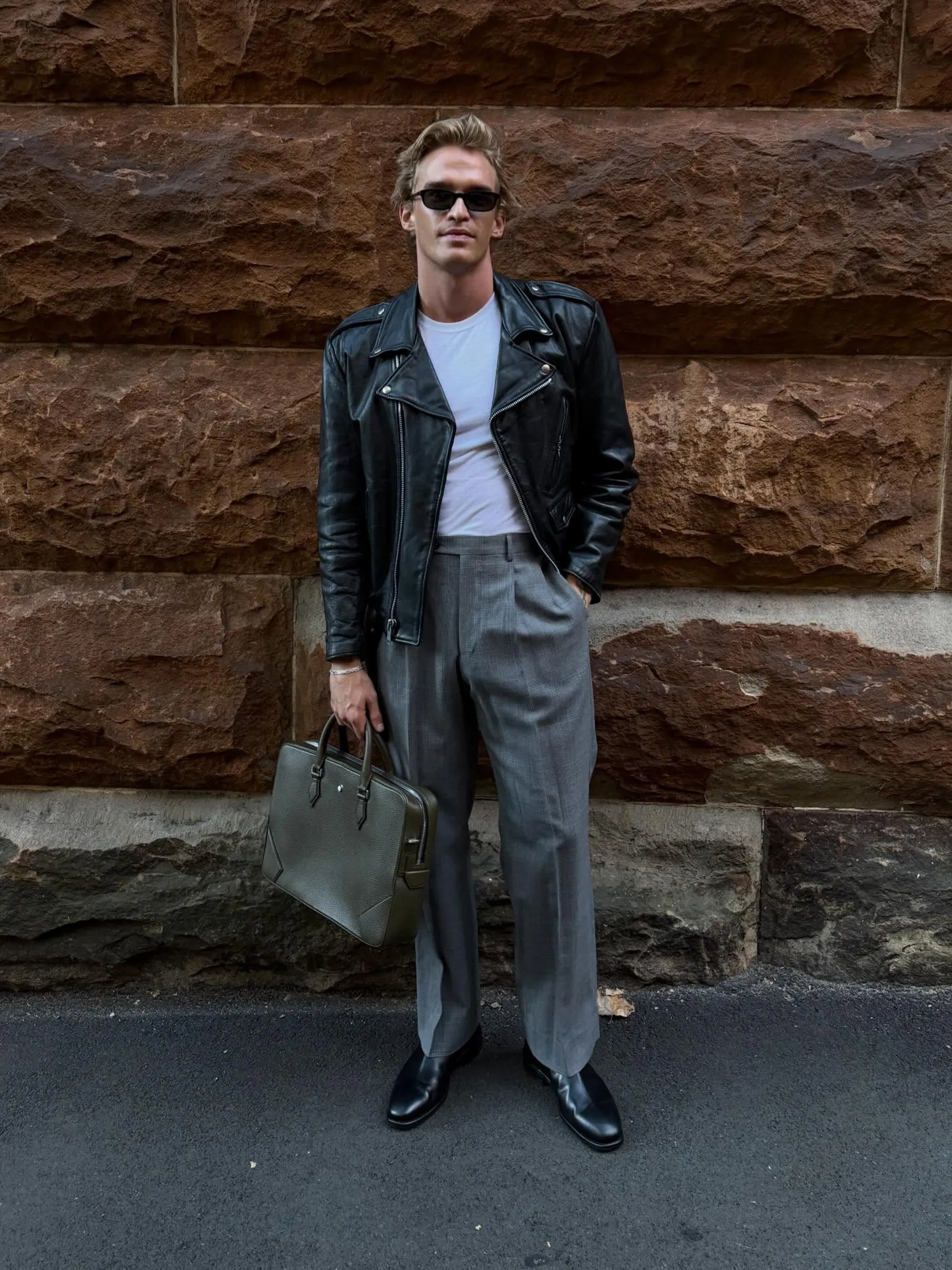 Cody Simpson in a black leather jacket, white t-shirt, gray trousers, and black shoes, holding a green briefcase.