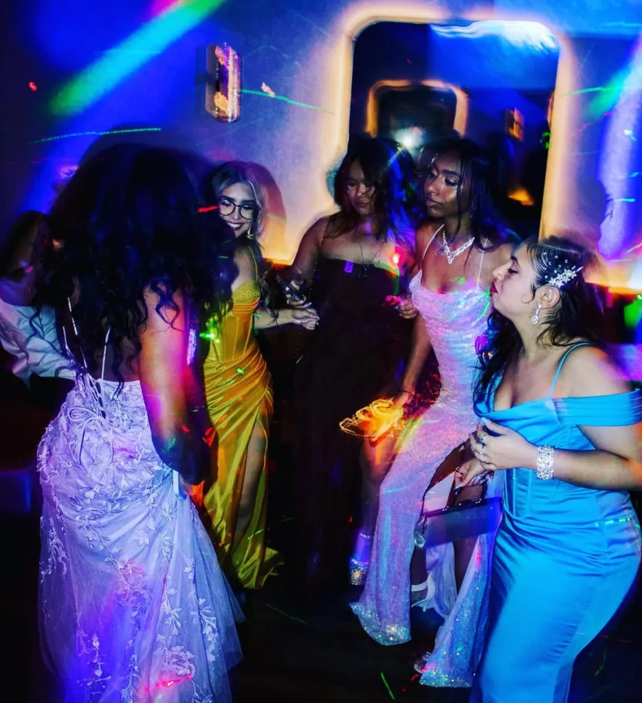 Five young women dancing in colorful prom dresses under party lights.