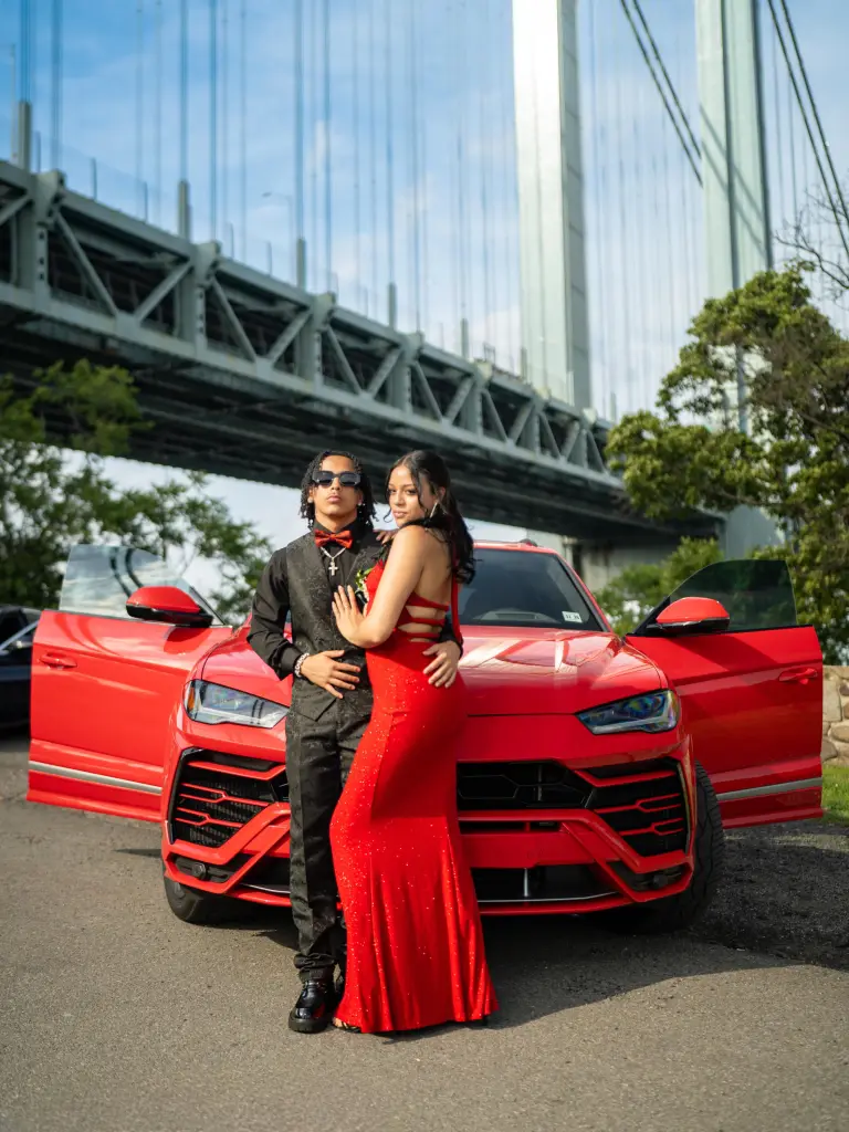 Two people posing in front of a red SUV, with a large bridge in the background.