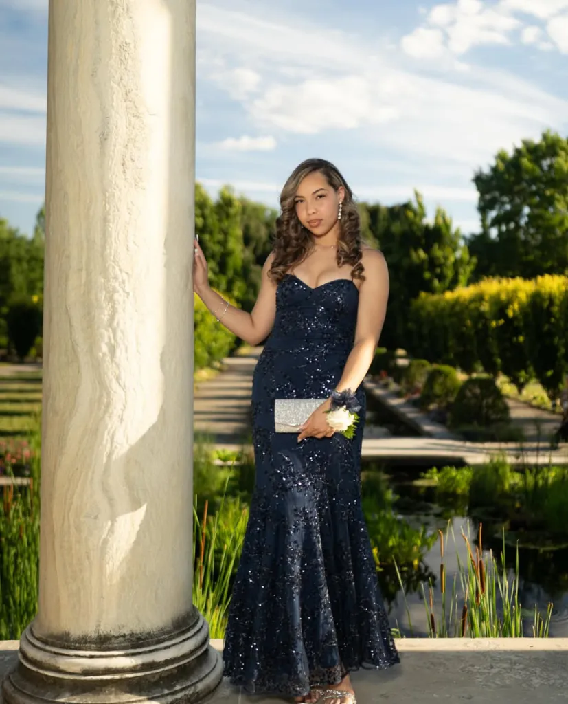 Young woman posing in a navy blue sequined gown next to a column outdoors.