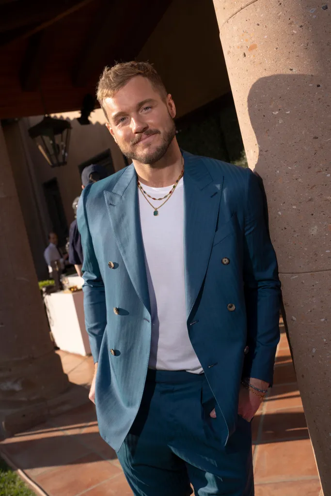 Colton Underwood in a teal suit jacket and pants, leaning against a stone column.