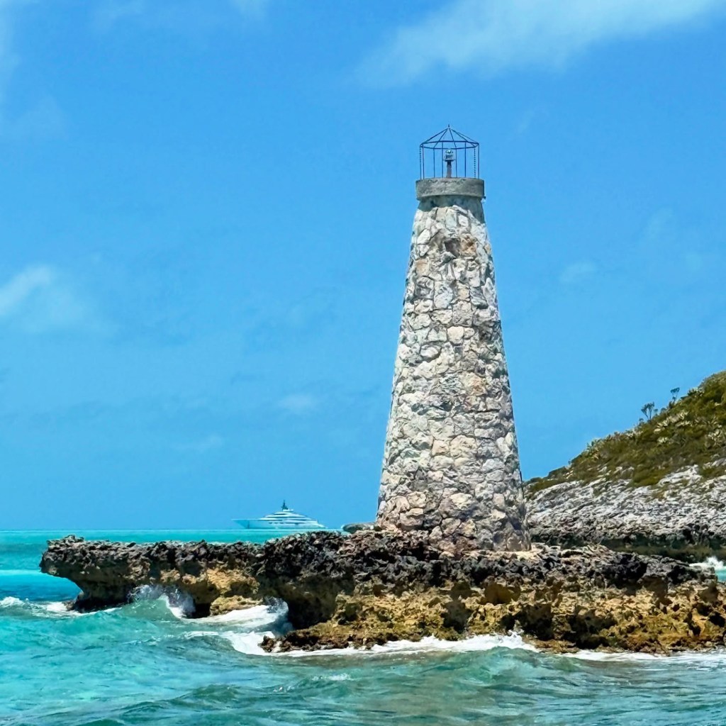 Stone lighthouse on a rocky Bahamian island with a yacht in the distance.