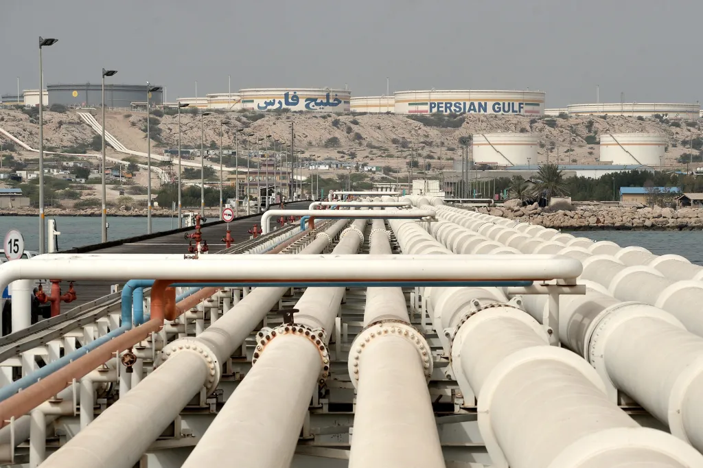 Numerous pipes extending from the foreground into the background, leading to large storage tanks on a rocky hill by the water.