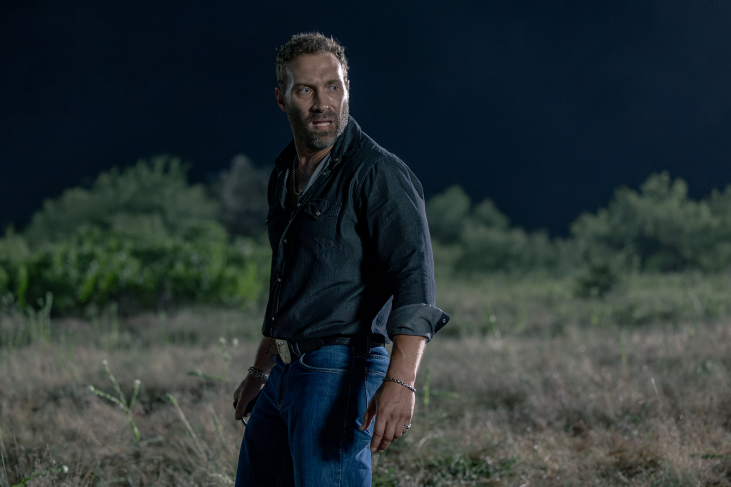 Jai Courtney as Rob-Will Jackson, looking fearful and dressed in a dark shirt and blue jeans, standing in a field at night.