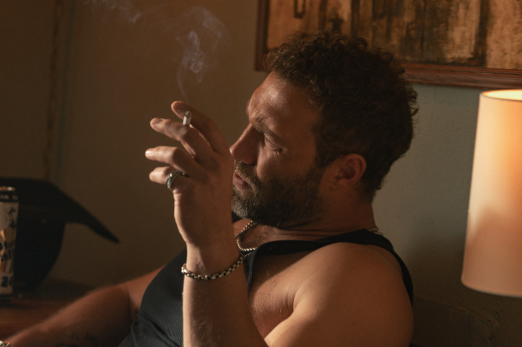 Jai Courtney as Rob-Will, a man with a beard and curly hair, smoking a cigarette while wearing a black tank top and a silver chain.