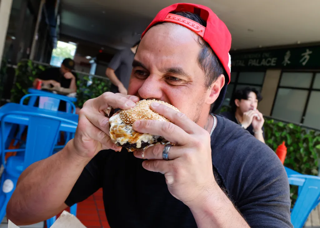 A man in a red hat takes a large bite out of a messy burger, with cheese and sauce dripping.