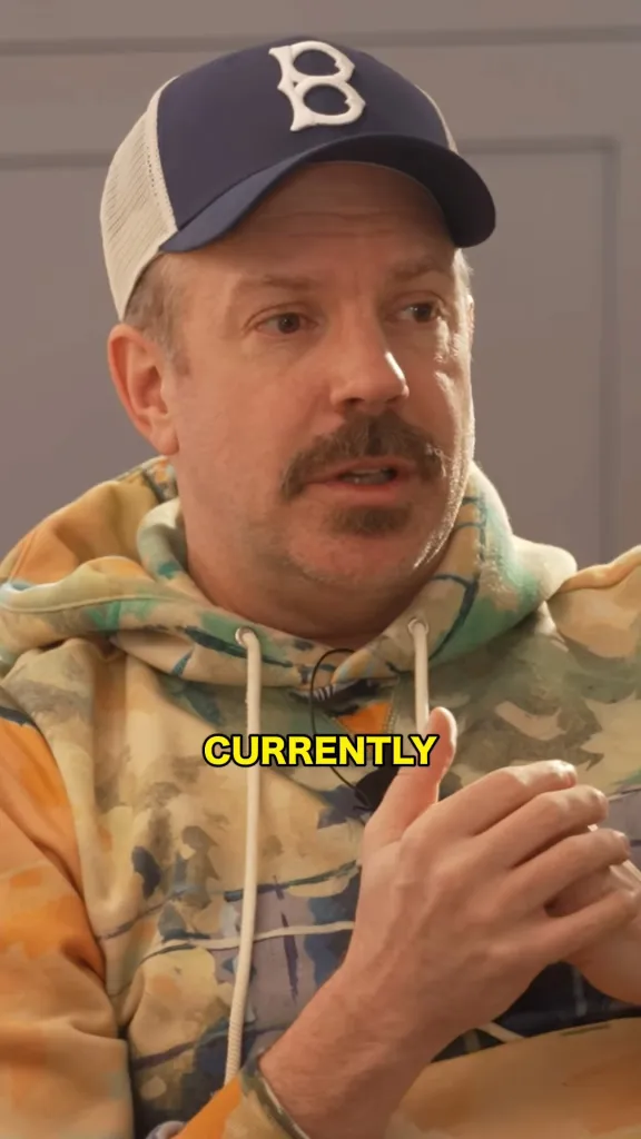 Jason Sudeikis in a blue and white baseball cap and tie-dye hoodie, with the word "CURRENTLY" superimposed.