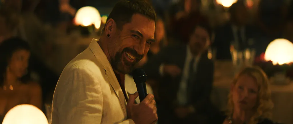 Javier Bardem smiles while holding a microphone at a dimly lit event.