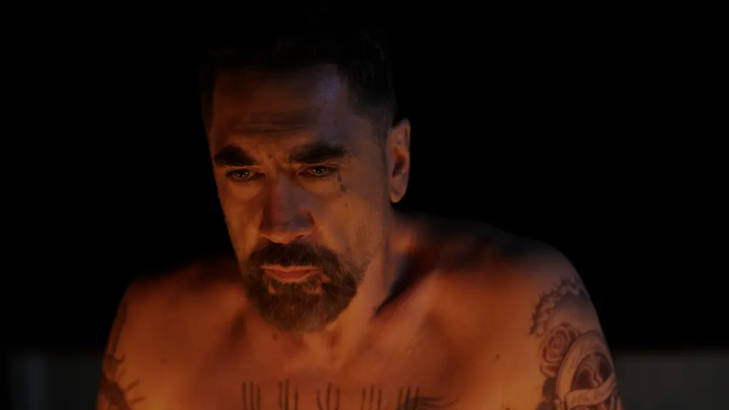 Javier Bardem as Max Cady, shirtless with tattoos, looking down in a dimly lit scene.