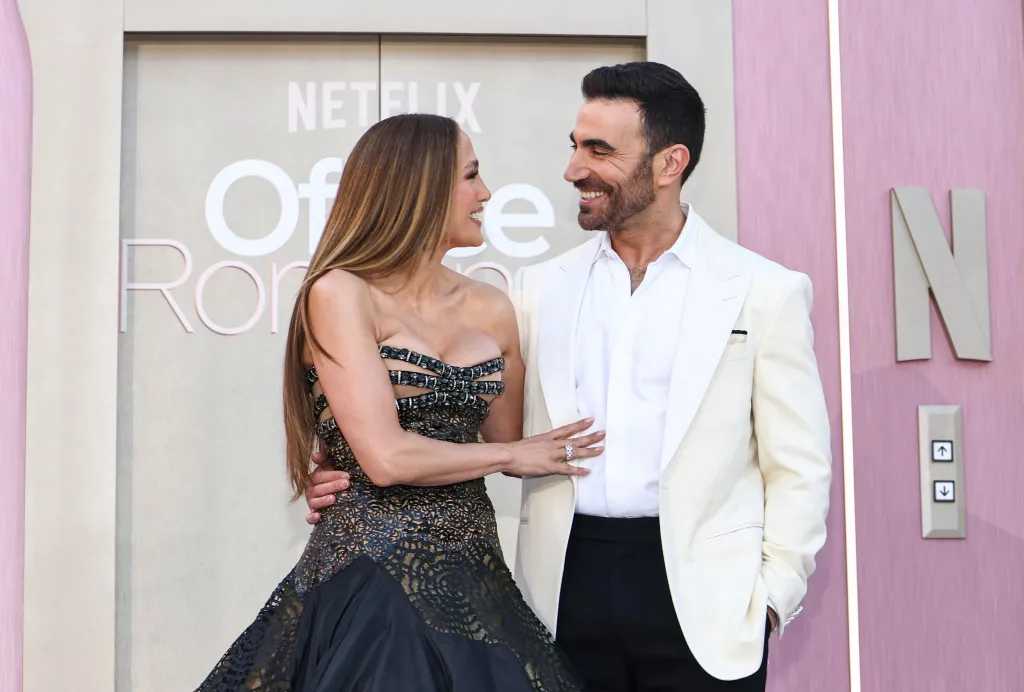 Jennifer Lopez in a black dress and Brett Goldstein in a white suit smile at each other at the "Office Romance" Film World Premiere.