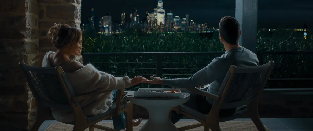 Jennifer Lopez as Jackie Cruz and Brett Goldstein as Daniel Blanchflower sharing a box of candy while sitting on a balcony overlooking a city skyline at night.