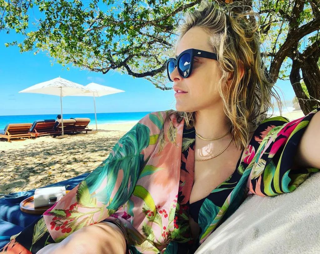 Jenny Mollen wearing a floral kimono and sunglasses on a beach with white umbrellas and the ocean in the background.