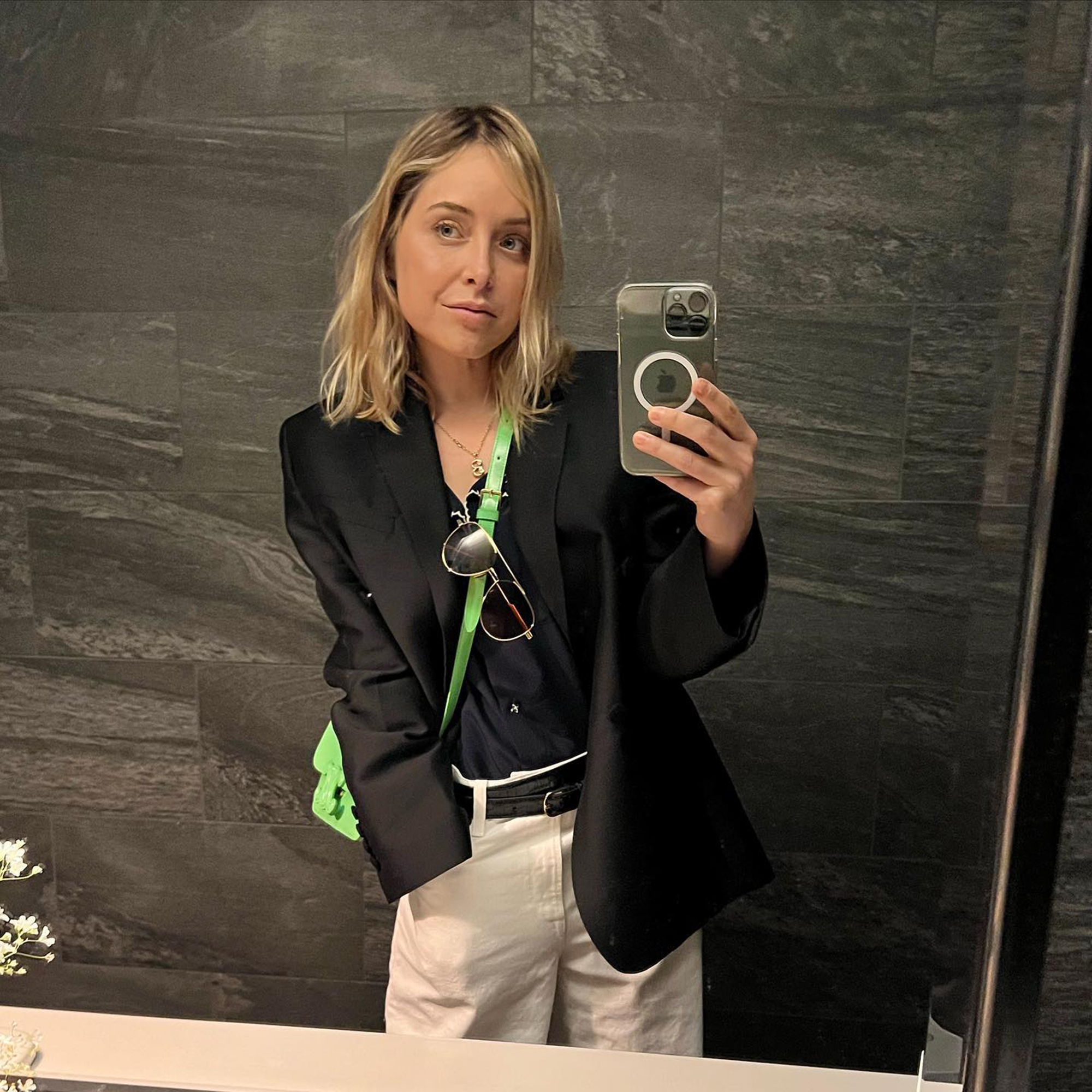 Jenny Mollen takes a selfie in a mirror, dressed in a black blazer, white shorts, and a neon green strap.