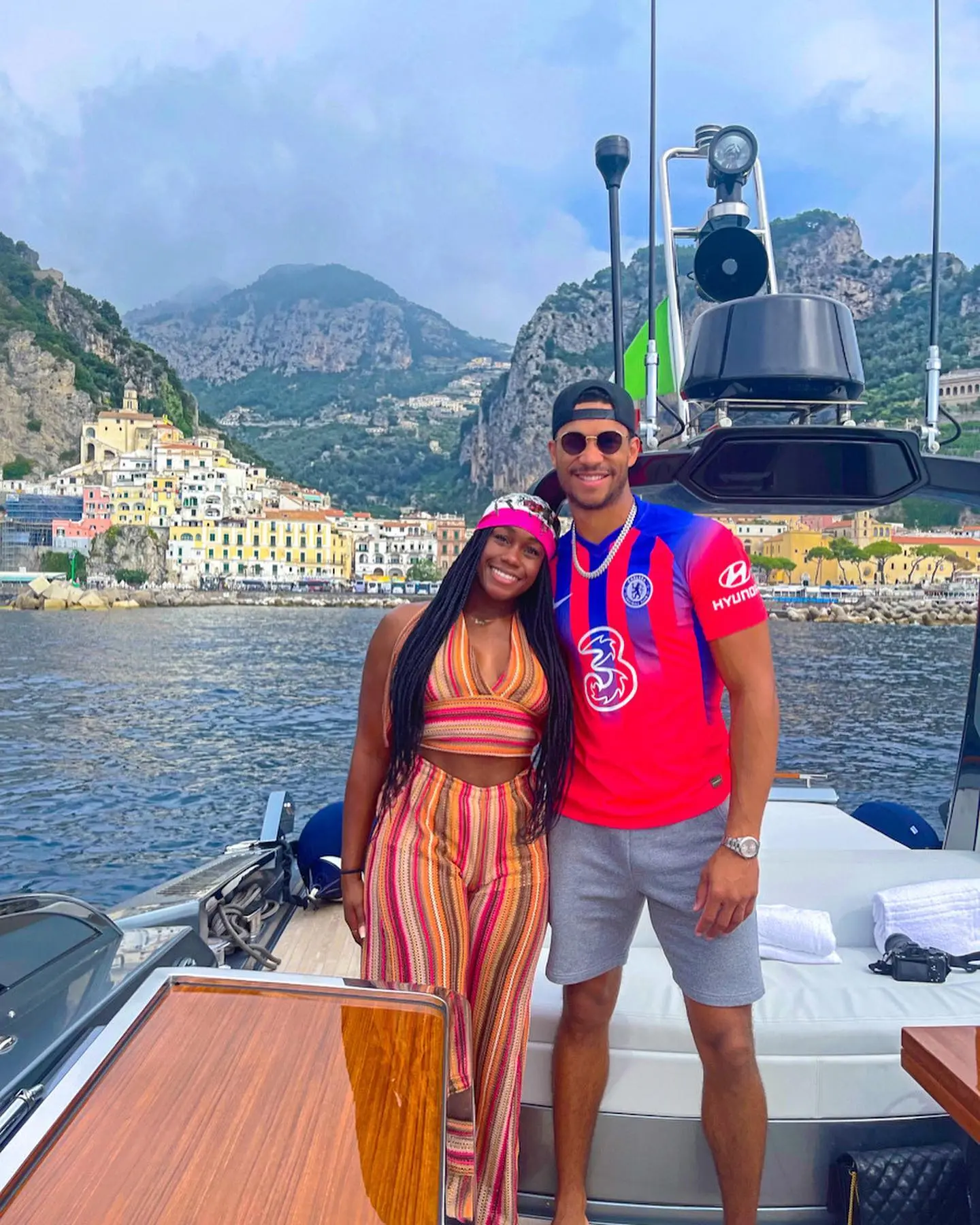 Josh Hart and Shannon posing on a boat with a scenic Italian coastal town in the background.