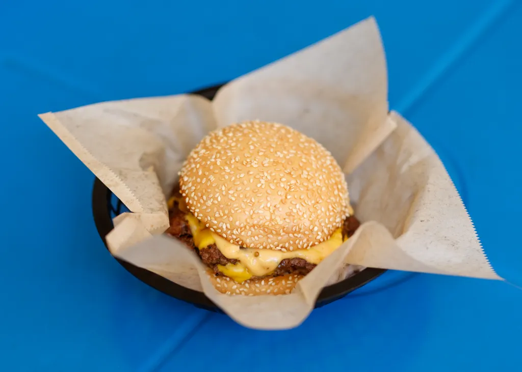 A double Amboy burger with sesame seed bun and melted cheese, served in a black basket lined with paper on a blue table.