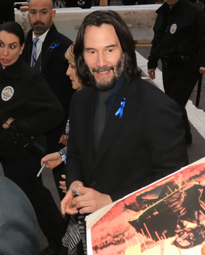 Keanu Reeves smiling and signing an autograph with a blue ribbon pinned to his suit.