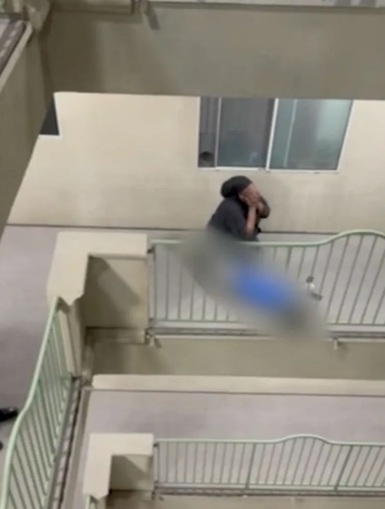 A person in a black shirt and head covering leans over a balcony railing, looking down with a distressed expression.