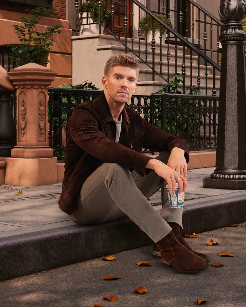 Kyle Cooke sitting on a sidewalk with a coffee cup, looking at the camera.