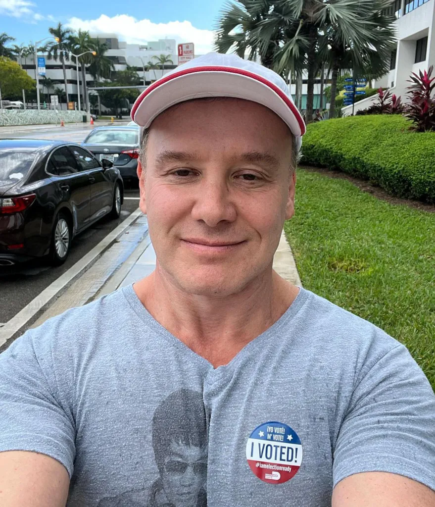 Lenny Hochstein wearing a gray t-shirt with an "I Voted!" sticker, a white hat with red trim, and standing outdoors on a sidewalk next to a street with cars parked.
