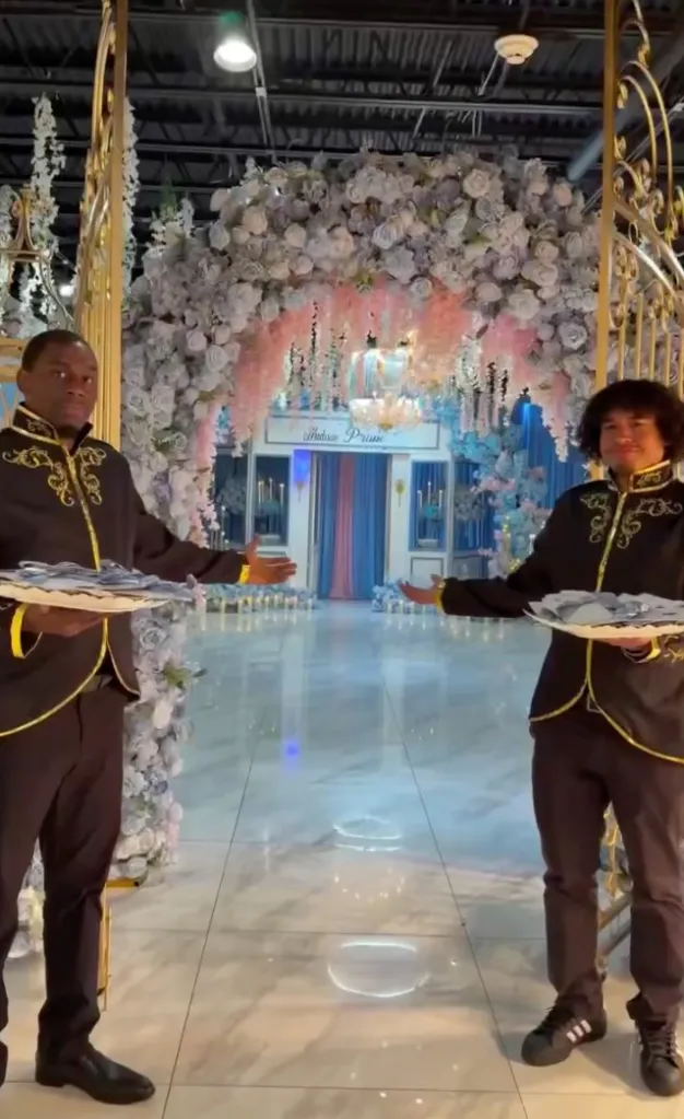 Two men in matching black and gold formal wear holding trays of party favors, standing in a flower-decorated archway.