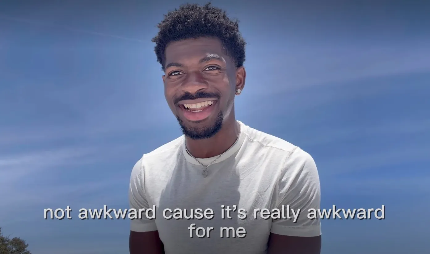 Lil Nas X smiling, wearing a white t-shirt and silver heart necklace, with text overlay