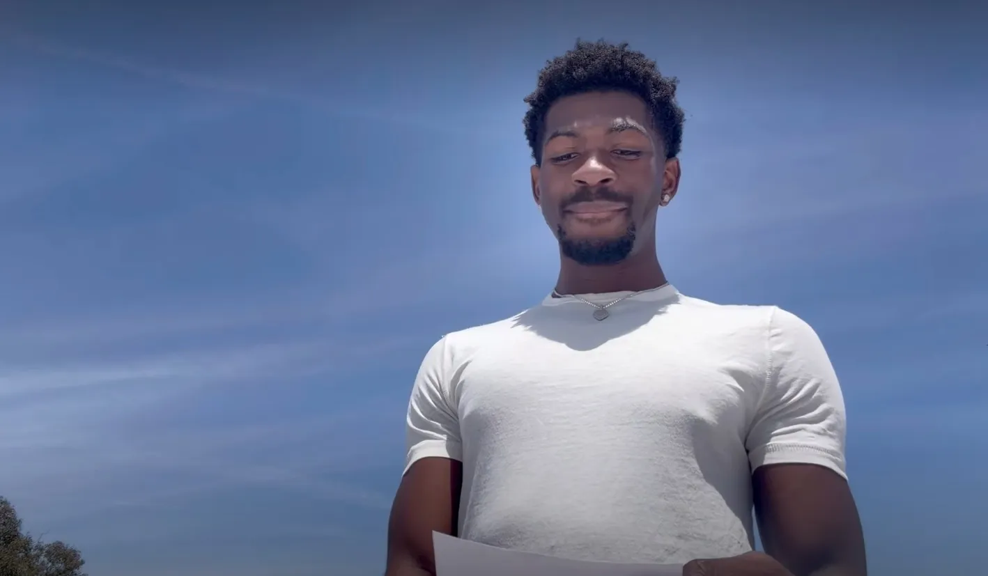 Lil Nas X wearing a white t-shirt and diamond earrings, looking down and holding a piece of paper.