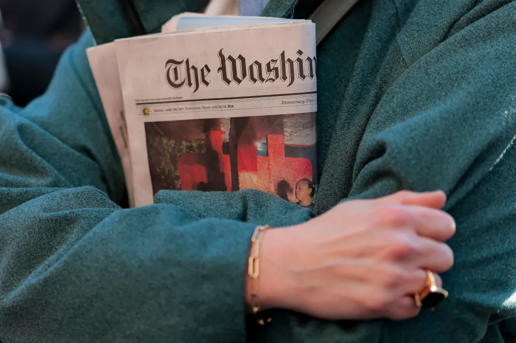 A person in a green coat holding a copy of The Washington Post newspaper.