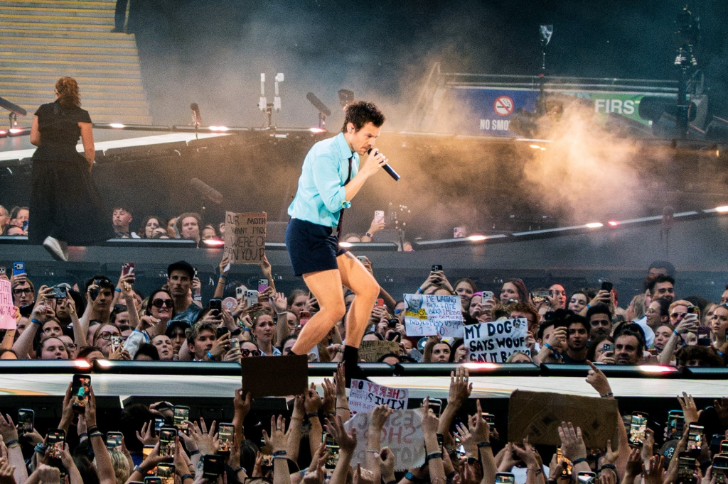 Harry Styles performing on stage at Wembley Stadium, surrounded by a crowd of fans holding up phones and signs.