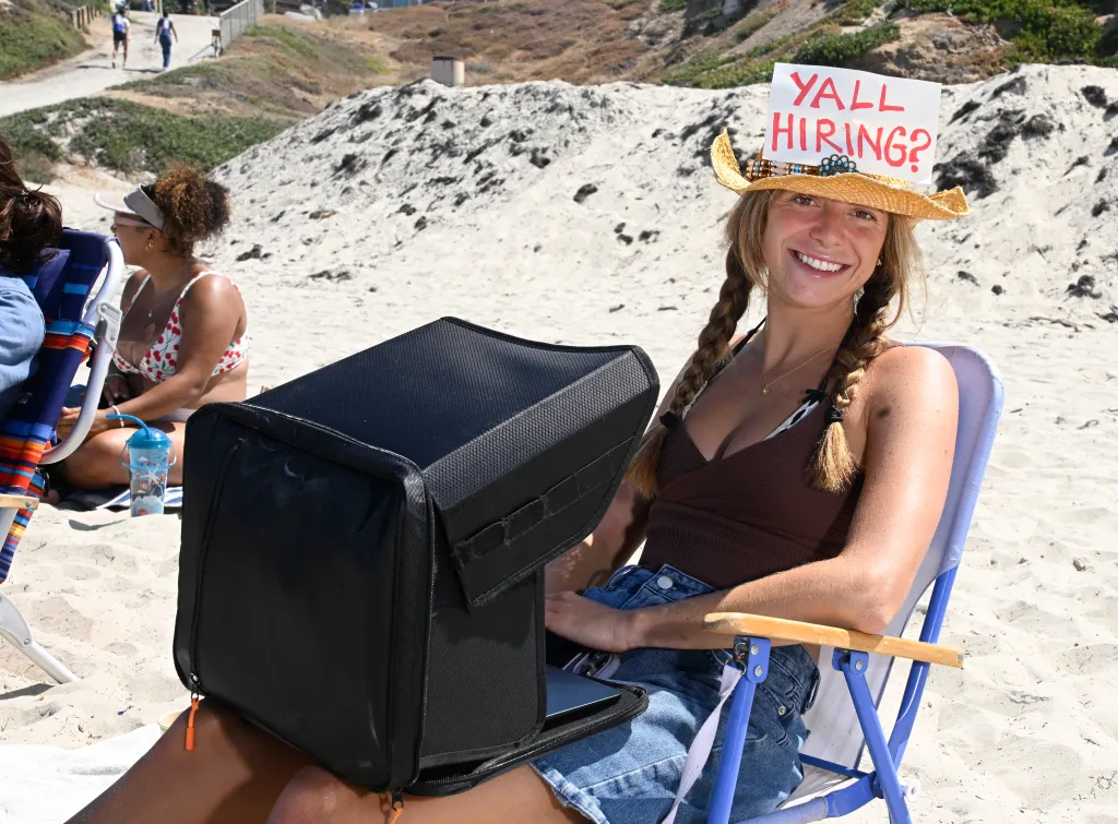 Maddy Singer works on her laptop at the beach in Pacific Beach, wearing a straw hat with a sign that says "YALL HIRING?".