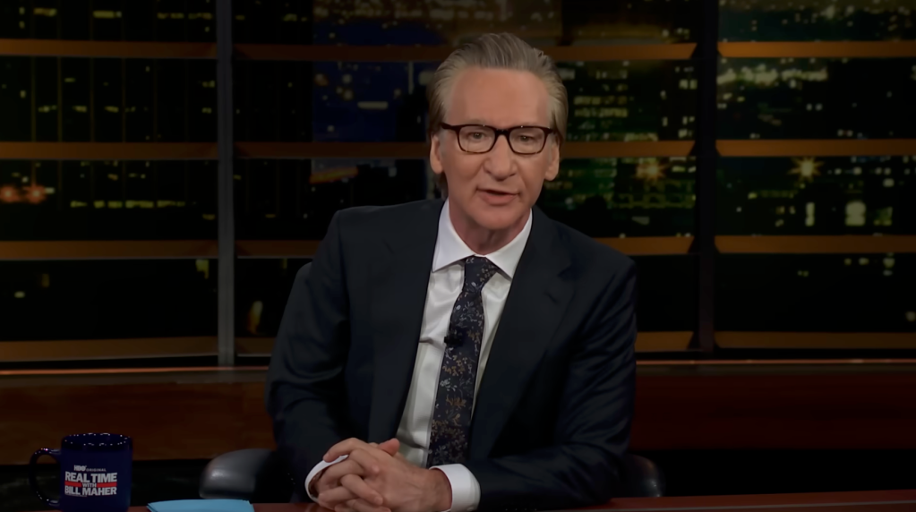Bill Maher sits on a set of "Real Time with Bill Maher" with a city skyline in the background.